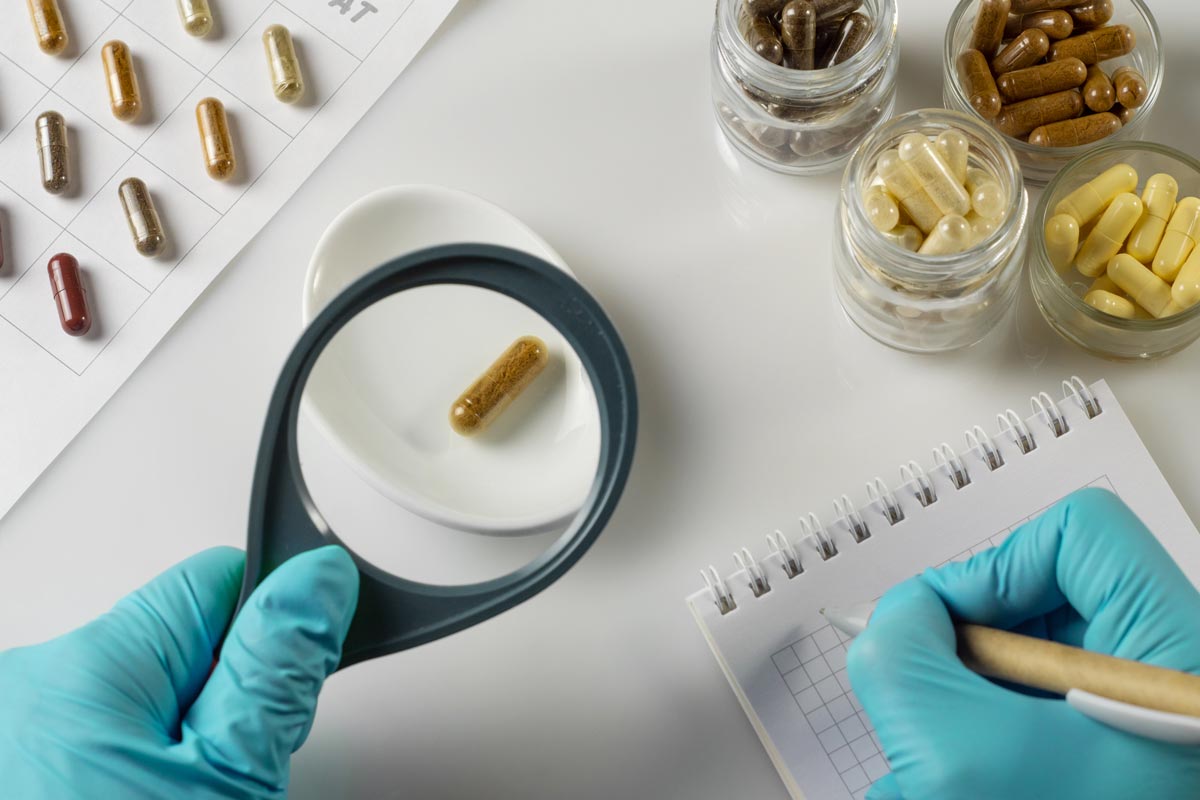 Hand of a pharmacist holds a capsule with dietary supplements and magnifying glass on the background of jars of ingredients in laboratory
