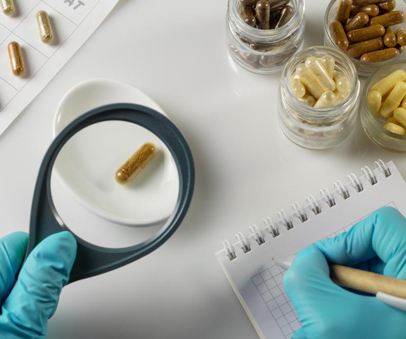 Hand of a pharmacist holds a capsule with dietary supplements and magnifying glass on the background of jars of ingredients in laboratory