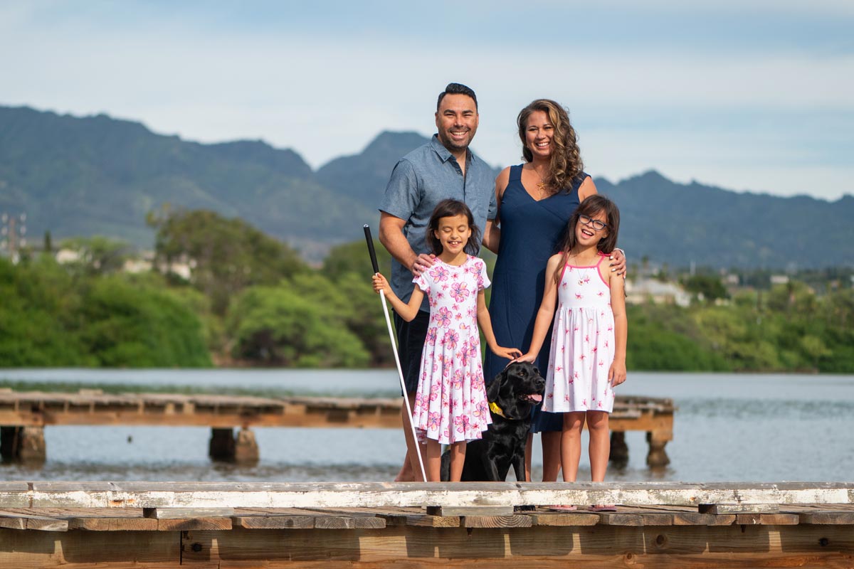 a dad, mom, two daughters and a dog stand on a dock with mountains in the background
