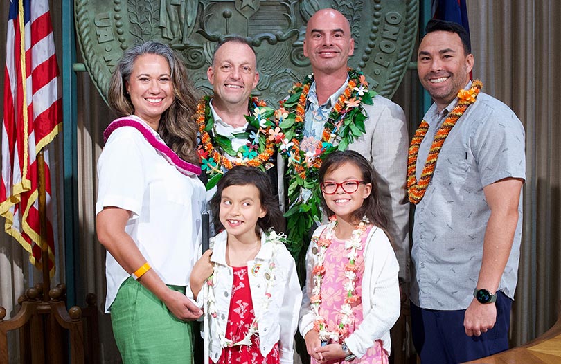 Group shot of family with young children with governor and trust leader in front of Hawaii state seal.
