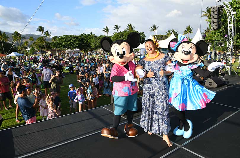 Woman wearing muumuu and lei on stage with Mickey and Minnie Mouse with a crowd of families in the audience.
