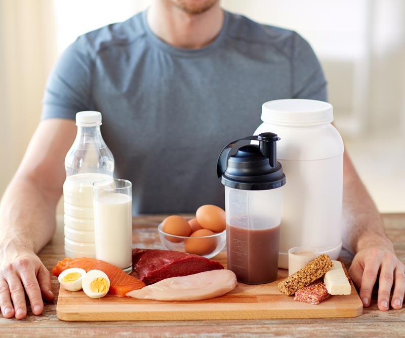 a man sits in front of a wooden tray stacked with milk, chicken, fish, steak, eggs and protein supplements