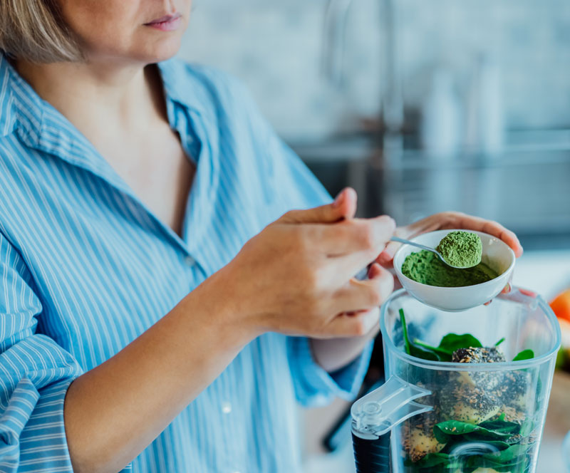 woman scooping greens powders into a blender full of fresh fruits and vegetables