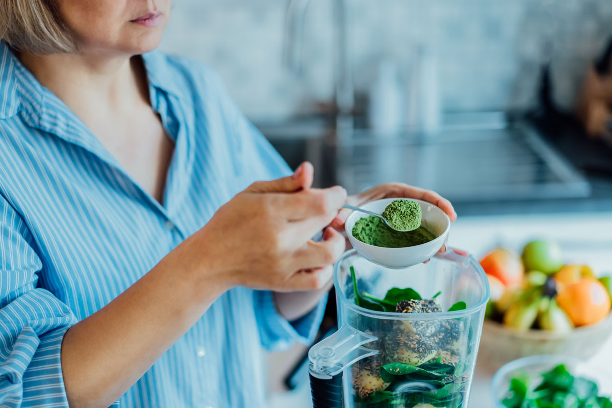 woman scooping greens powders into a blender full of fresh fruits and vegetables