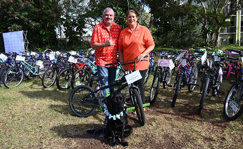 Two adults and a service dog pose in front of a row of kid's bicycles to be given away.
