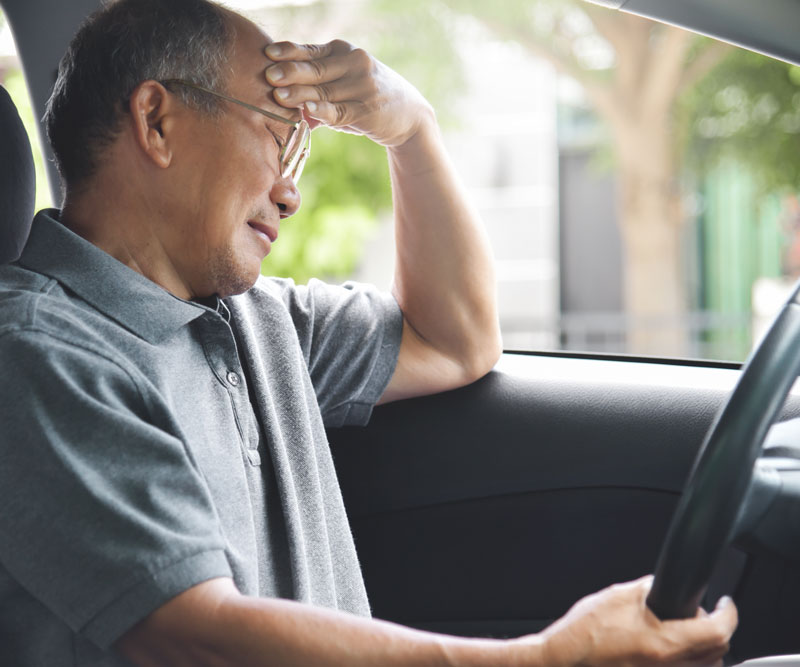 older man holds head while sitting in the driver's seat of a car