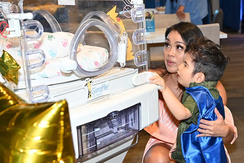 Mother with young son, wearing blue cape, looking at an incubator.