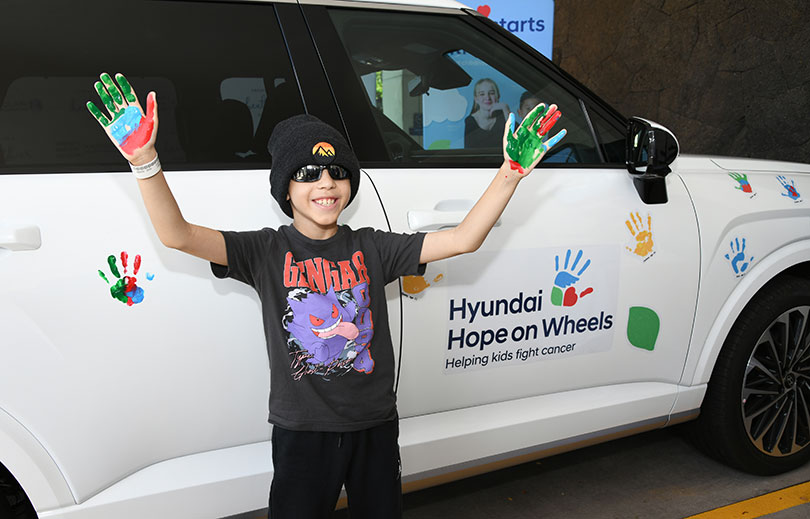 Young boy in beanie and sunglasses with upraised palms painted colorfully standing in front of the Hyundai Hope on Wheels vehicle, decorated with colorful handprints.