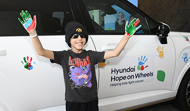 Young boy in beanie and sunglasses with upraised palms painted colorfully standing in front of the Hyundai Hope on Wheels vehicle, decorated with colorful handprints.