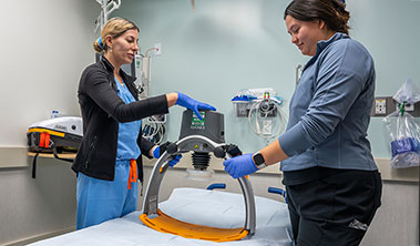 Two medical professionals holding an automated CPR machine in a medical exam room.